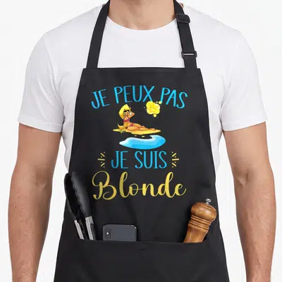 Tablier de cuisine noir avec une femme blonde en bikini sous le soleil et un texte humoristique.