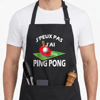 Tablier de cuisine noir avec une raquette de ping pong rouge, une balle blanche et le texte J'PEUX PAS J'AI PING PONG en blanc.