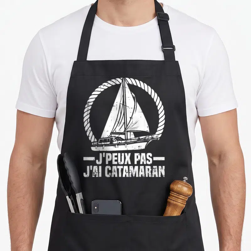 Tablier de cuisine noir avec illustration de catamaran et texte J'peux pas j'ai catamaran capitaine en blanc.