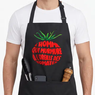 Tablier de cuisine noir avec une illustration de tomate rouge formant le texte l'homme qui murmure à l'oreille des tomates et des feuilles vertes.