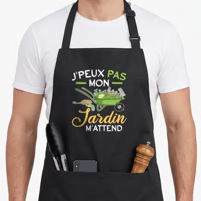 Tablier de cuisine noir avec une brouette verte remplie d'outils et le texte J'peux pas mon Jardin m'attend en typographie colorée.