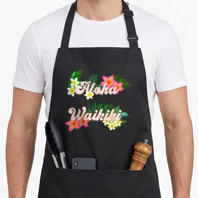Tablier de cuisine noir avec inscription Aloha Waikiki entourée de fleurs tropicales roses et blanches ainsi que de feuilles de palmier vertes.