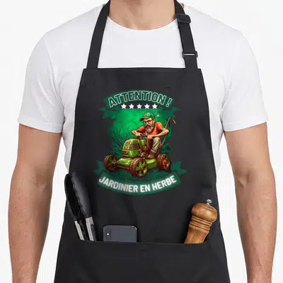 Tablier de cuisine noir avec illustration d'un homme barbu sur une tondeuse verte et des bandes textuelles.