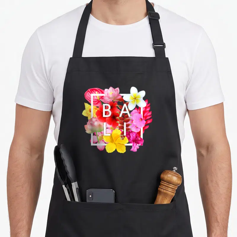 Tablier de cuisine noir avec composition florale tropicale et texte blanc BALI au centre.