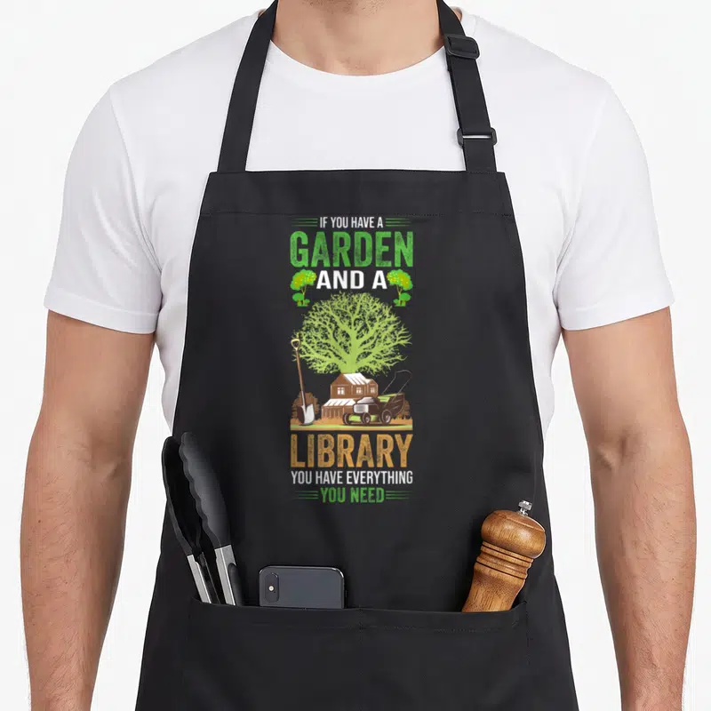 Tablier de cuisine noir avec illustration d'un grand arbre, d'une maison, d'une pelle et d'un taille-haie sous une inscription bilingue sur le jardinage et la lecture.