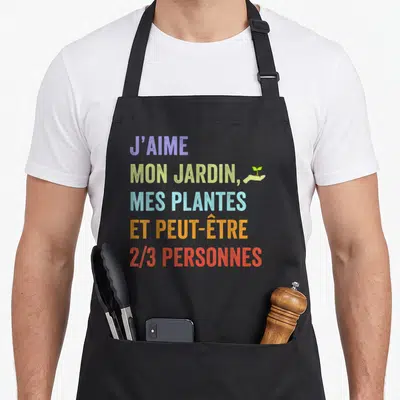 Tablier de cuisine noir avec texte coloré J'aime mon jardin mes plantes et peut-être 2/3 personnes accompagné d'une main tenant une graine.