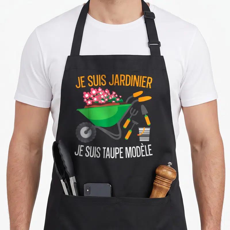 Tablier de cuisine noir avec illustration de brouette verte remplie de fleurs roses et d'outils de jardinage aux poignées orange.