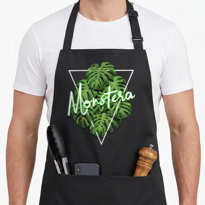 Tablier de cuisine noir avec une illustration de feuilles de Monstera Deliciosa et le texte Monstera en lettres blanches stylisées.