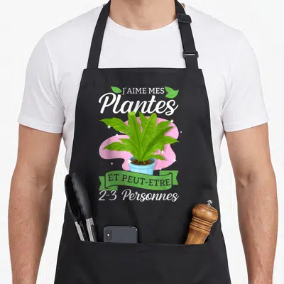 Tablier de cuisine noir avec illustration d'une plante verte en pot et texte humoristique sur fond rose.