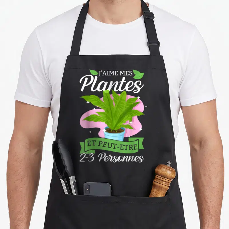 Tablier de cuisine noir avec illustration d'une plante verte en pot et texte humoristique sur fond rose.