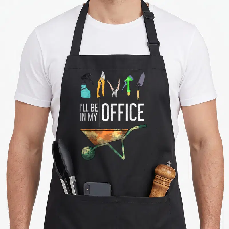 Tablier de cuisine noir avec inscription I'll Be In My Office, outils de jardinage et brouette rouillée colorée.