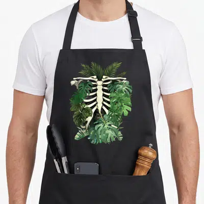 Tablier de cuisine noir avec illustration d'un squelette de thorax entouré de feuilles de monstera vertes et détaillées.