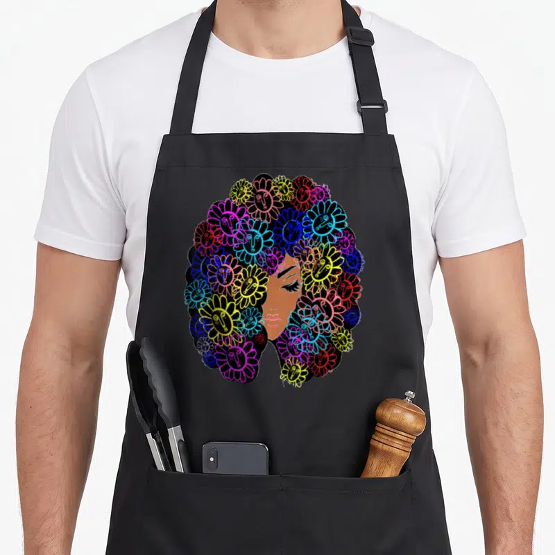 Tablier de cuisine noir avec illustration d'un visage afro entouré de marguerites colorées aux visages imprimés.