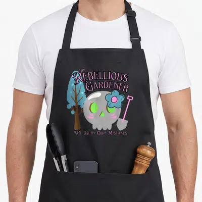 Tablier de cuisine noir avec un crâne rose, une fleur bleue et la mention The Rebellious Gardener en lettres stylisées.