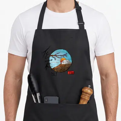 Tablier de cuisine noir avec illustration circulaire du mont Fuji, fleurs de cerisier et symboles kanji sur fond bleu ciel.