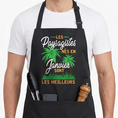 Tablier de cuisine noir avec inscription Les Paysagistes nés en janvier sont les meilleurs et deux palmiers verts sur fond de verdure.