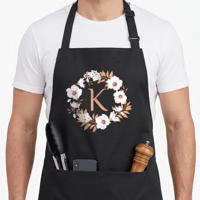 Tablier de cuisine noir avec une couronne de fleurs roses et blanches entourant la lettre K rose poudré au centre.
