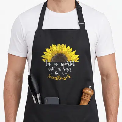 Tablier de cuisine noir avec un grand tournesol jaune et le texte In a world full of roses be a sunflower en calligraphie blanche et dorée.