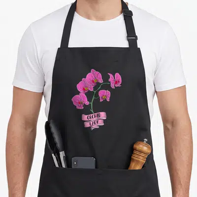 Tablier de cuisine noir avec une illustration colorée d'orchidées roses et un ruban indiquant ORCHID LADY