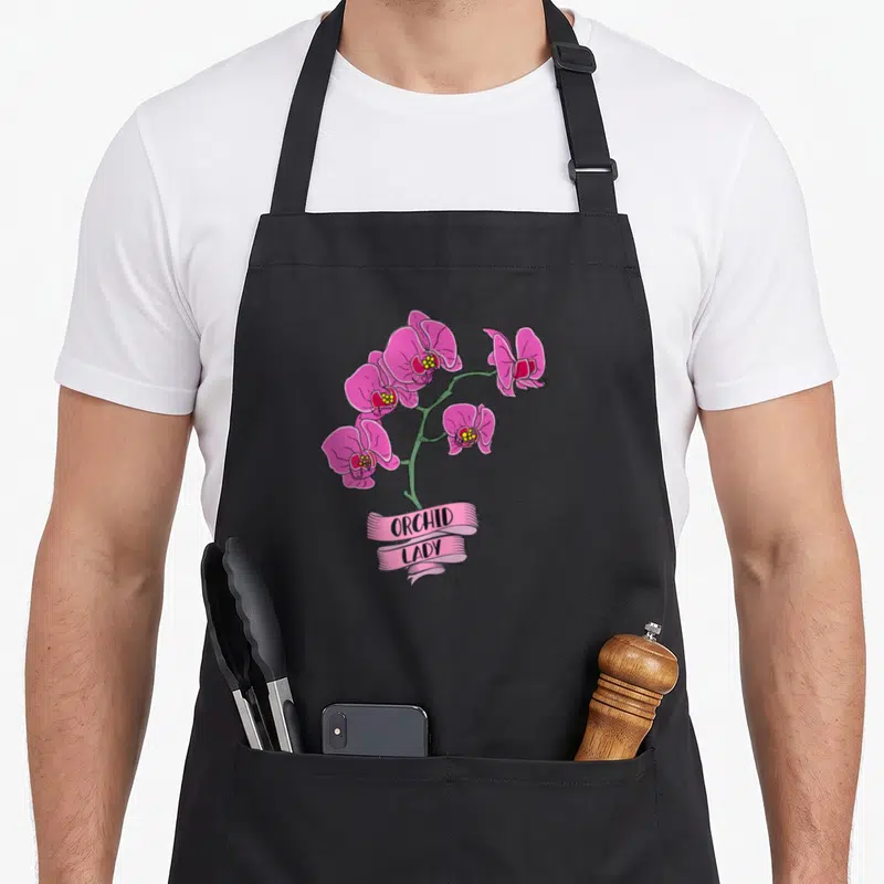 Tablier de cuisine noir avec une illustration colorée d'orchidées roses et un ruban indiquant ORCHID LADY