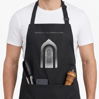 Tablier de cuisine noir avec une arche blanche géométrique rayonnante et le texte The Battle at Garden's Gate en haut.