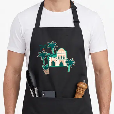 Tablier de cuisine noir avec illustration d'une mosquée, de palmiers verts et d'un cactus en pot rose.