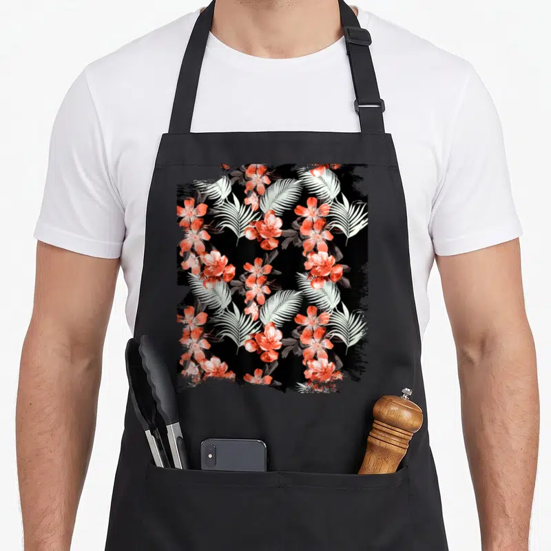 Tablier de cuisine noir avec motifs de feuilles de palmier blanches et fleurs rouges vives disposées en motif répétitif.