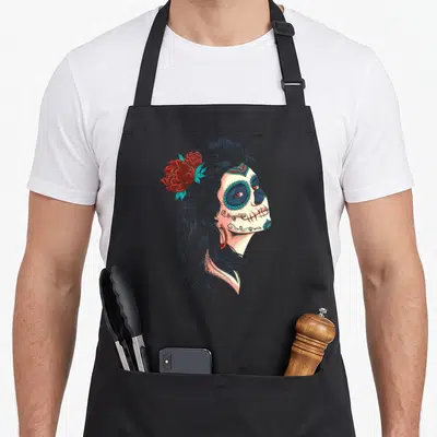Tablier de cuisine noir avec illustration latérale d'une Calavera Catrina coiffée de fleurs rouges et de motifs turquoise.
