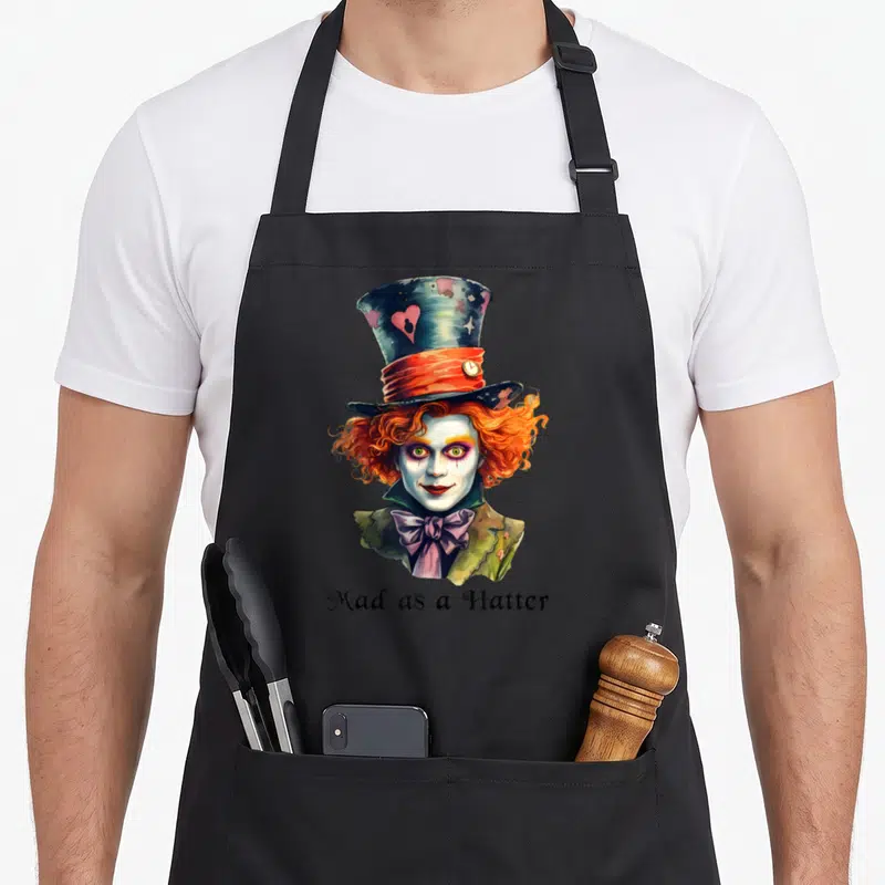 Tablier de cuisine noir avec illustration du Chapelier fou aux cheveux orange et texte Mad as a Hatter en bas.