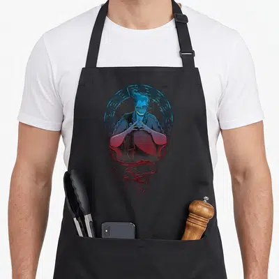 Tablier de cuisine noir avec portrait stylisé de Hades encadré de motifs géométriques grecs en bleu et rouge.