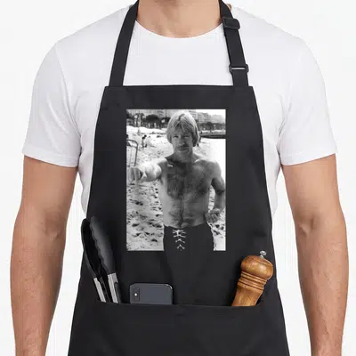 Tablier de cuisine noir avec une illustration monochrome d'un homme musclé pointant vers l'avant sur un fond de plage.