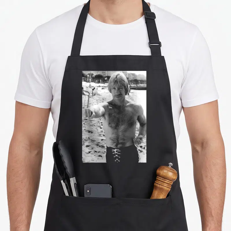 Tablier de cuisine noir avec une illustration monochrome d'un homme musclé pointant vers l'avant sur un fond de plage.