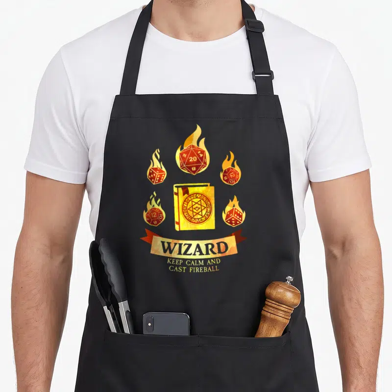 Tablier de cuisine noir avec un livre de magicien doré, des dés enflammés et le mot WIZARD sur une bannière.