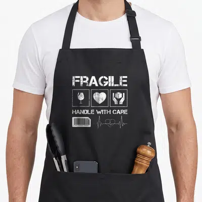 Tablier de cuisine noir avec inscription Fragile Handle With Care et pictogrammes blancs stylisés.