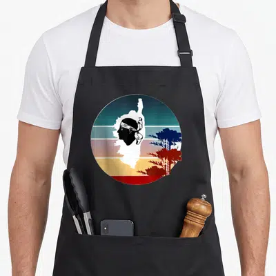 Tablier de cuisine noir avec une illustration circulaire représentant la carte de Corse, un profil au bandana et des arbres sur fond de bandes colorées vintage.