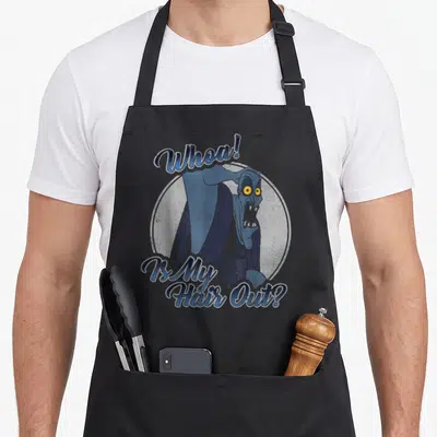 Tablier de cuisine noir avec une illustration bleue du personnage Hades et le texte Whoa Is My Hair Out imprimé en typographie stylisée.