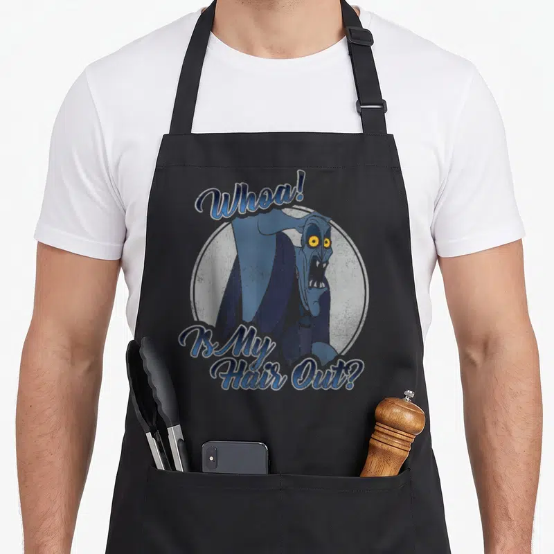 Tablier de cuisine noir avec une illustration bleue du personnage Hades et le texte Whoa Is My Hair Out imprimé en typographie stylisée.