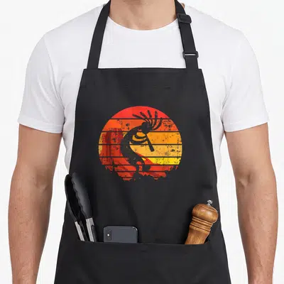 Tablier de cuisine noir avec illustration circulaire Kokopelli orange et jaune sur fond désaturé