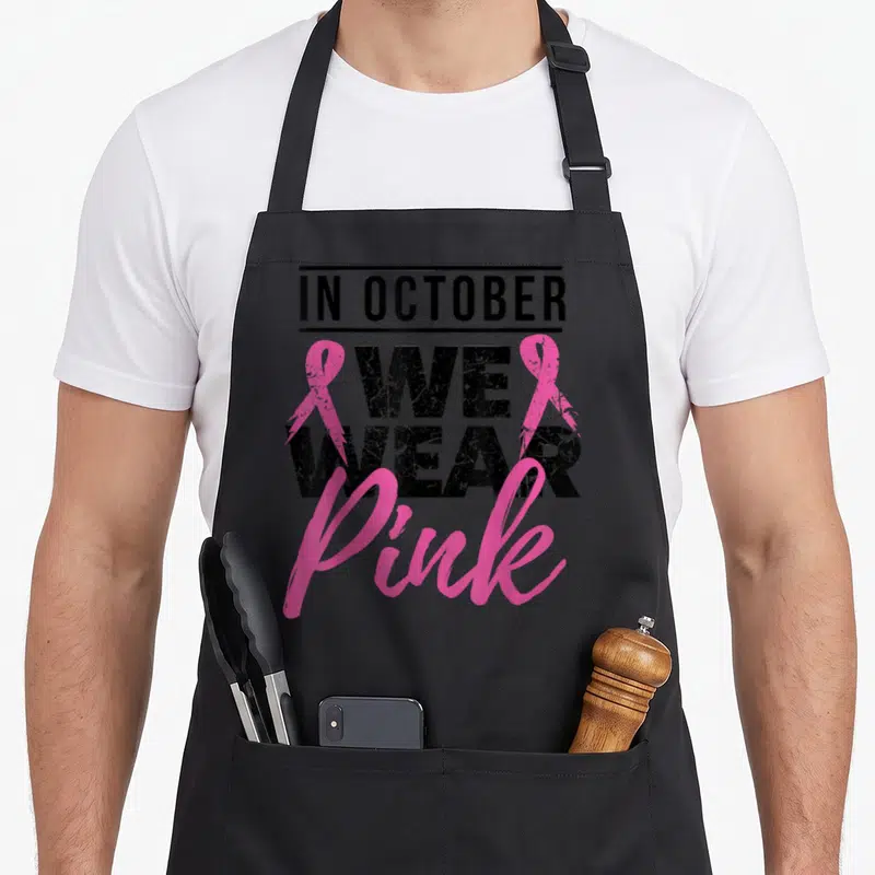 Tablier de cuisine noir avec l'inscription In October Nous Portons Pink et deux rubans roses stylisés.