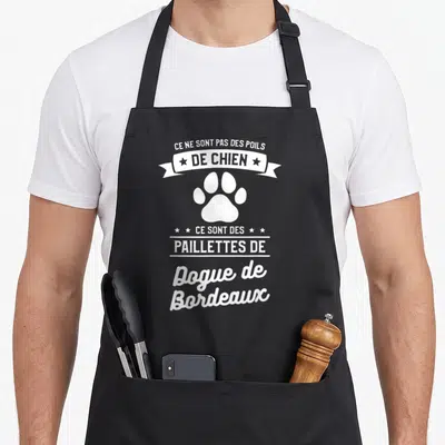 Tablier de cuisine noir avec inscription humoristique sur un fond gris uni et une patte de chien blanche.