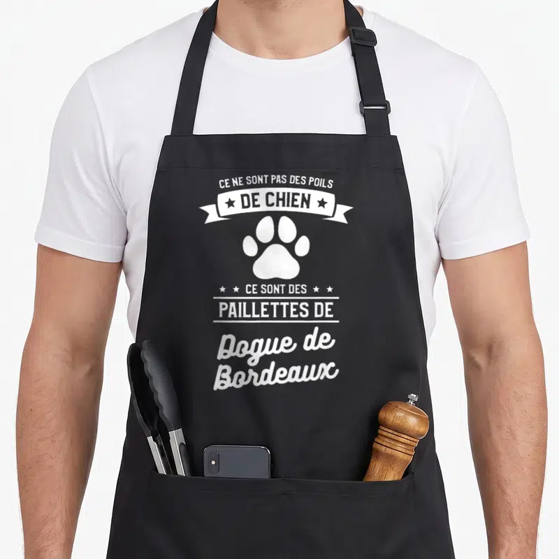Tablier de cuisine noir avec inscription humoristique sur un fond gris uni et une patte de chien blanche.