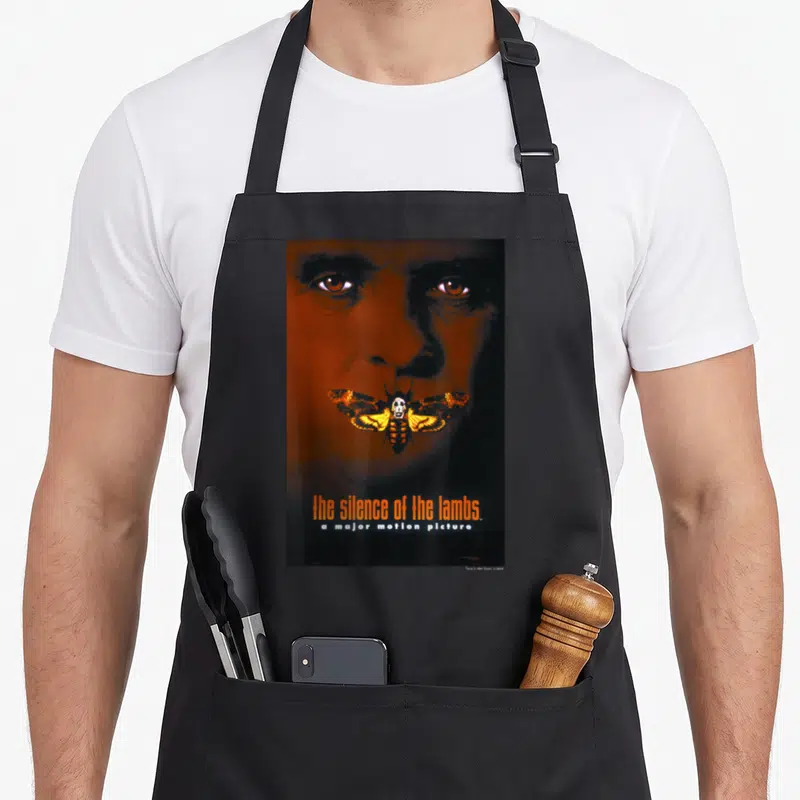 Tablier de cuisine noir avec illustration d'un visage aux yeux orangés et un papillon portant un masque blanc au centre.
