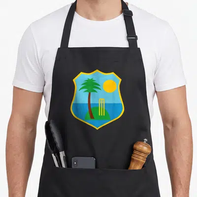 Tablier de cuisine noir avec un blason tropical illustrant un palmier et des wickets de cricket sous un soleil jaune.