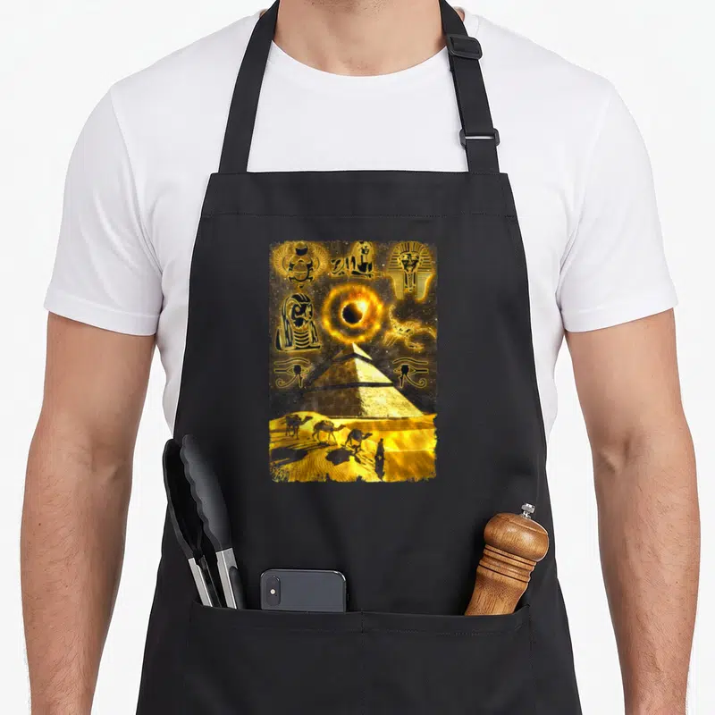 Tablier de cuisine noir avec illustration dorée de pyramides, œil d'Horus et chameaux dans le désert.