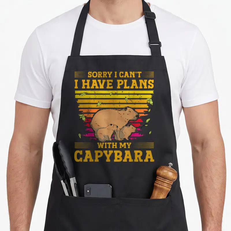 Tablier de cuisine noir avec illustration de trois capybaras et texte jaune sur fond coloré dégradé.