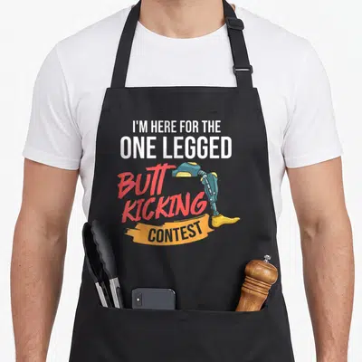 Tablier de cuisine noir avec une prothèse de jambe et le texte I'M HERE FOR THE ONE LEGGED BUTT KICKING CONTEST en lettres colorées.