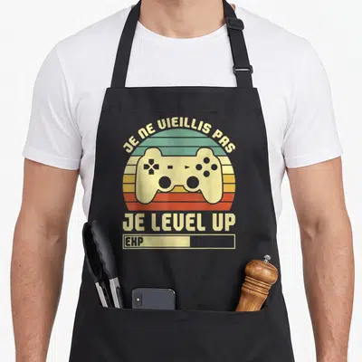 Tablier de cuisine noir avec une manette de jeu vidéo et le texte Je ne vieillis pas je level up en style rétro.
