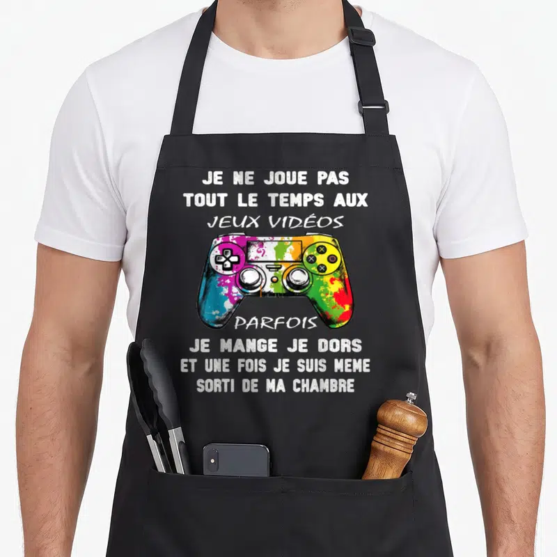 Tablier de cuisine noir avec manette de jeu multicolore et texte blanc humoristique sur fond gris.