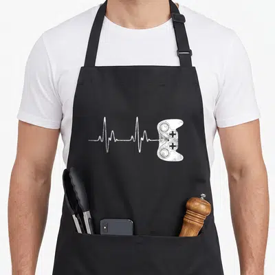 Tablier de cuisine noir avec un motif blanc représentant une ligne de rythme cardiaque reliée à une manette de jeu vidéo stylisée.