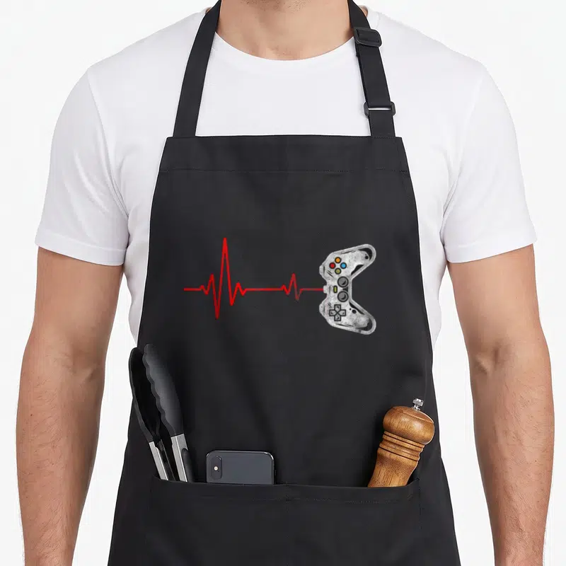 Tablier de cuisine noir avec une illustration de manette de jeu et une ligne de rythme cardiaque rouge.